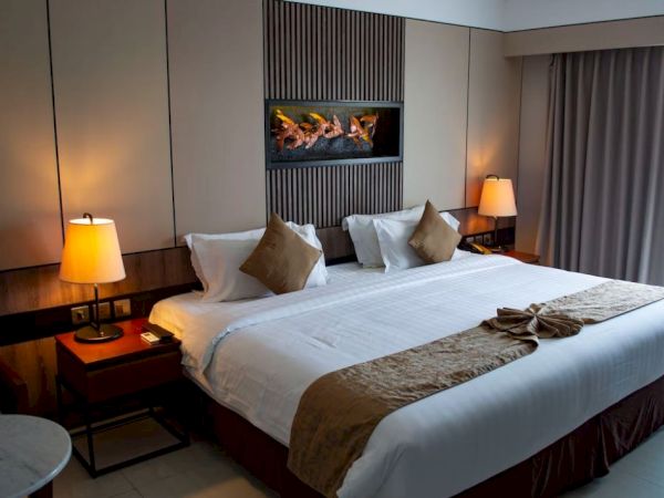 A cozy hotel room with a large bed, decorative pillows, warm lighting, wooden accents, and a wall-mounted faux fireplace.