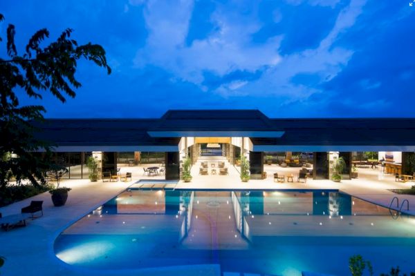 A luxurious outdoor pool area at dusk, featuring a modern building with open seating and ambient lighting under a partly cloudy sky.