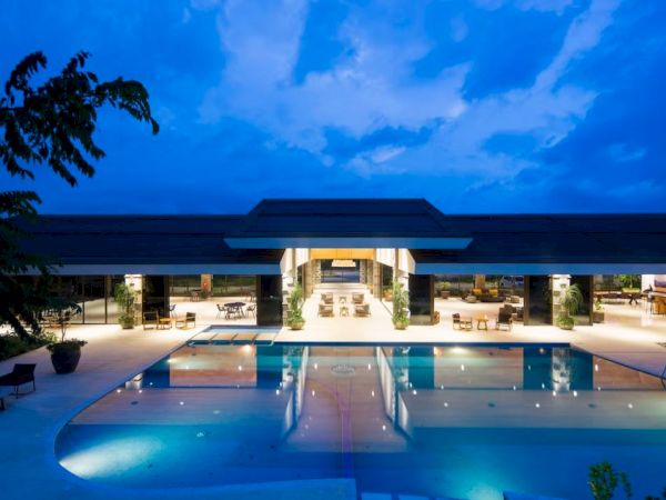 A luxurious outdoor pool area at dusk, featuring a modern building with open seating and ambient lighting under a partly cloudy sky.