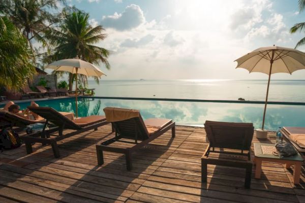 A tranquil poolside scene with lounge chairs, umbrellas, and palm trees, overlooking a calm sea under a partly cloudy sky.