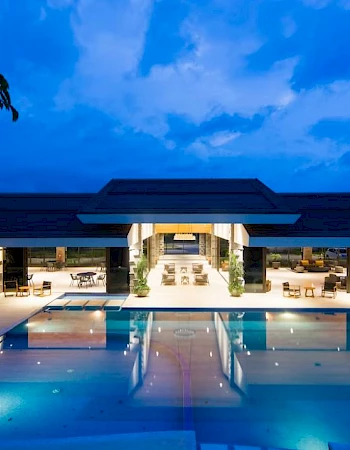 A luxurious modern building with an illuminated swimming pool at dusk, featuring open seating areas and a serene blue sky backdrop.