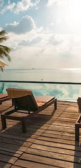 Poolside scene with wooden deck chairs, umbrellas, palm trees, and a serene ocean view under a cloudy sky with sunlight peeking through.