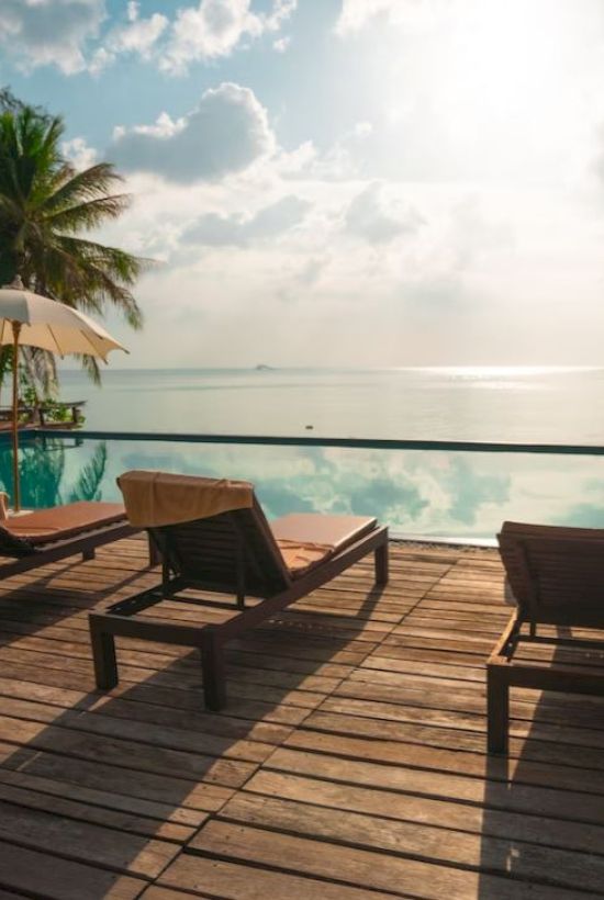 Lounge chairs and umbrellas by a tranquil pool, with palm trees and an ocean view under a partly cloudy sky at sunset.
