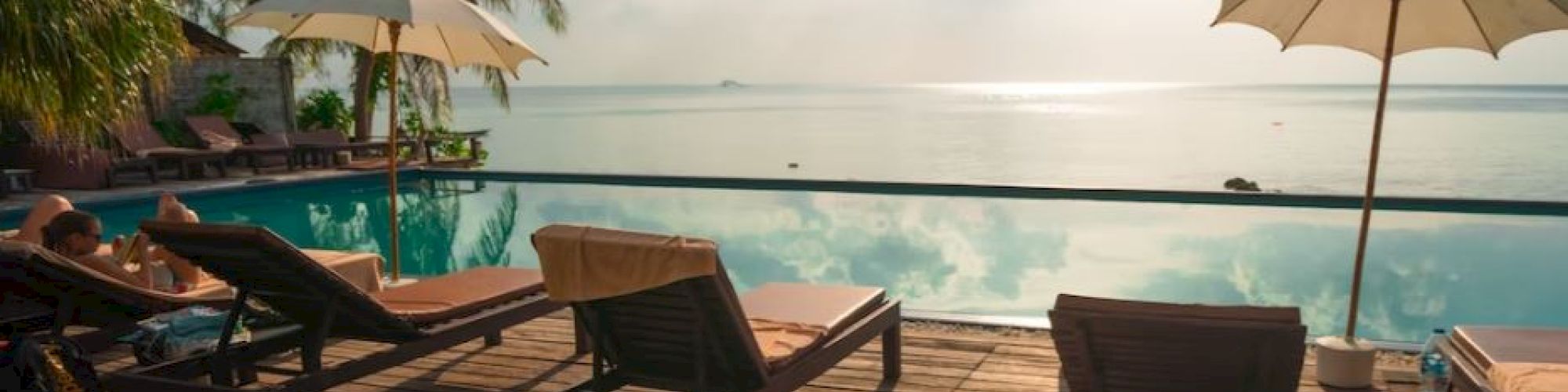 Lounge chairs and umbrellas by a pool, overlooking the ocean with palm trees and a bright sky in the background.