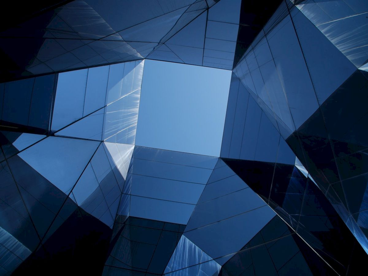 A geometric architectural view looking up, with reflective glass panels forming a symmetrical pattern against a clear blue sky.