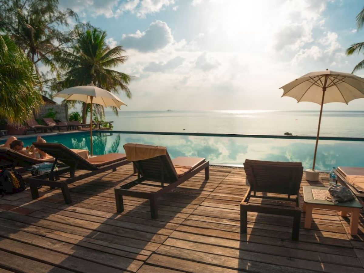 A serene poolside scene with lounge chairs, umbrellas, and palm trees overlooking a calm body of water under a sunlit sky.