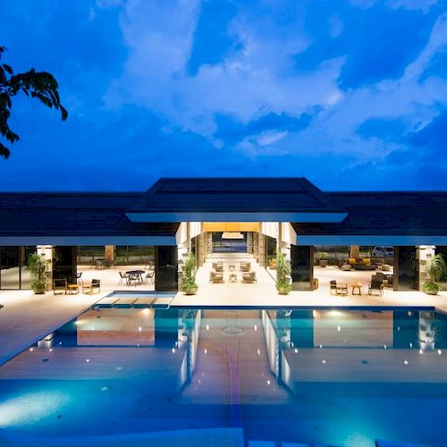 An illuminated modern building with a large pool in front, surrounded by lounge chairs, set against a blue twilight sky.