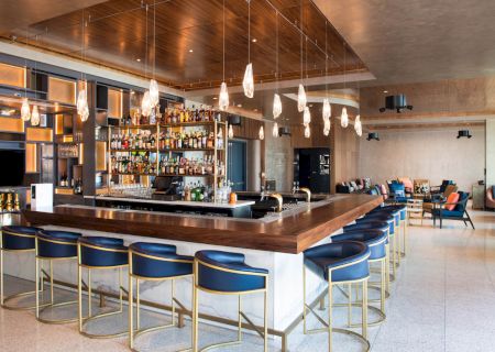 The image shows a modern bar with a wooden counter, blue stools, hanging lights, and shelves stocked with bottles, set in a spacious lounge.