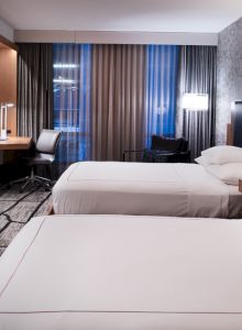 A modern hotel room with two beds, a TV, a desk, and large windows with curtains is pictured.