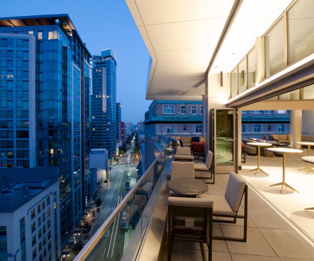 A modern balcony overlooking a city street, with tables and chairs, surrounded by tall buildings during dusk.