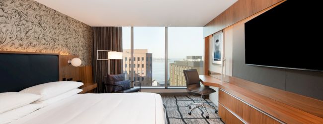 A modern hotel room with a large bed, TV, desk, chair, and window view of buildings and water in the background.
