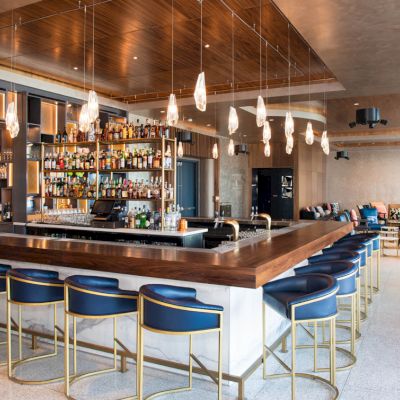 A modern bar with a wooden counter, hanging lights, and blue stools. Shelves with bottles line the back, with seating in the background.