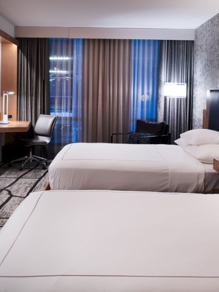 The image shows a modern hotel room with two double beds, a flat-screen TV, a desk, and stylish decor, featuring a patterned wall.