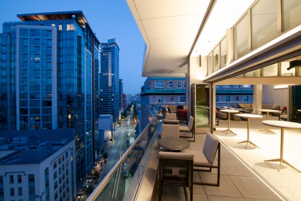 A modern urban balcony overlooking a city street with tall buildings, featuring seating and tables under a partially covered area.
