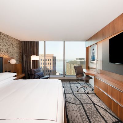 A modern hotel room with a large bed, desk, TV, and a window with a cityscape view. The decor includes wood accents and patterned walls.