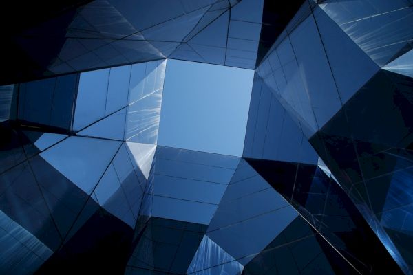 The image shows a view upward through reflective glass architecture, creating geometric patterns against a clear blue sky.