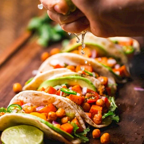 A hand squeezes lime over tacos filled with chickpeas, vegetables, and avocado on a wooden board.