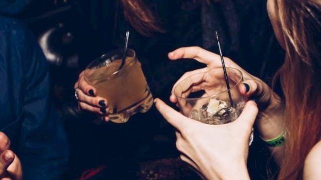 People are socializing, holding drinks, and laughing in what appears to be a lively, indoor setting.
