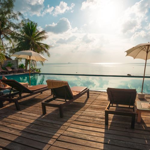 A serene poolside scene with lounge chairs, umbrellas, palm trees, and an ocean view under a partly cloudy sky and sunlight.