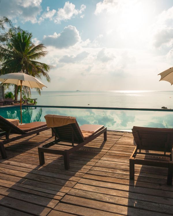 A serene poolside scene with lounge chairs, umbrellas, palm trees, and an ocean view under a partly cloudy sky and sunlight.