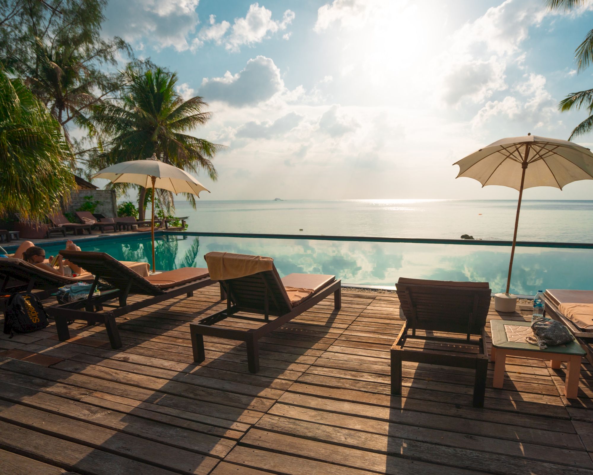 Chairs on a wooden deck facing an infinity pool with ocean views; palm trees and umbrellas create a tropical atmosphere.