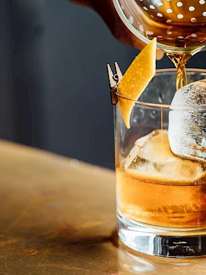 A cocktail being poured into a glass with ice and a citrus peel garnish on a wooden surface.