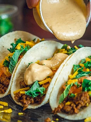 Three tacos with ground meat, lettuce, tomatoes, and cheese on a plate, topped with sauce being poured over them.