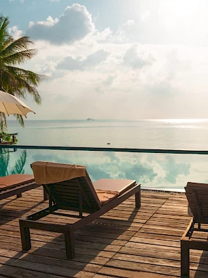 Lounge chairs and umbrellas on a wooden deck overlook an infinity pool, with palm trees and an ocean view under a partly cloudy sky.