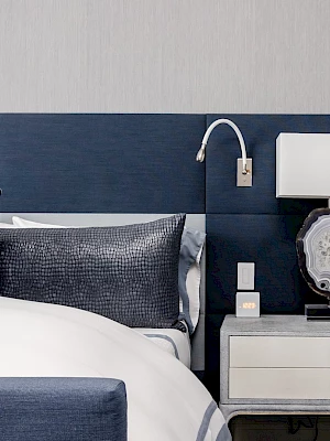 A bedroom with a navy headboard, bed, pillows, nightstand, lamp, and wall-mounted reading light.
