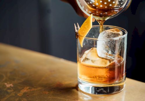 A drink being poured into a glass with ice and a lemon twist garnish, placed on a bar counter.