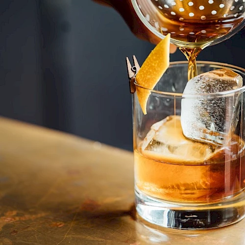 A drink being poured into a glass with ice and a lemon twist garnish, placed on a bar counter.