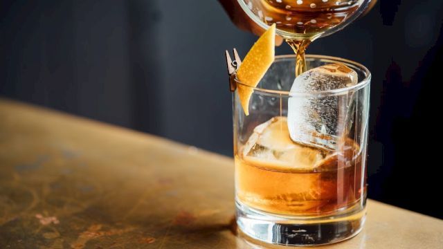 A drink being poured into a glass with ice and a lemon twist garnish, placed on a bar counter.