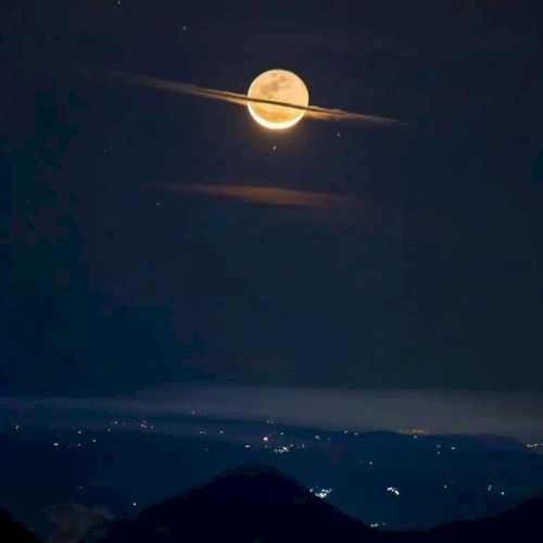 A full moon appears to have rings like Saturn due to passing clouds, set against a night sky over a landscape with twinkling lights.