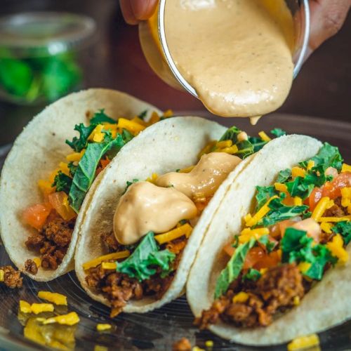 Three tacos with ground meat, cheese, lettuce, and tomatoes are on a plate, with sauce being poured over them.