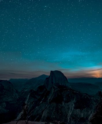 A starry night sky over a mountain landscape with a glowing horizon, silhouetted trees, and rugged terrain.