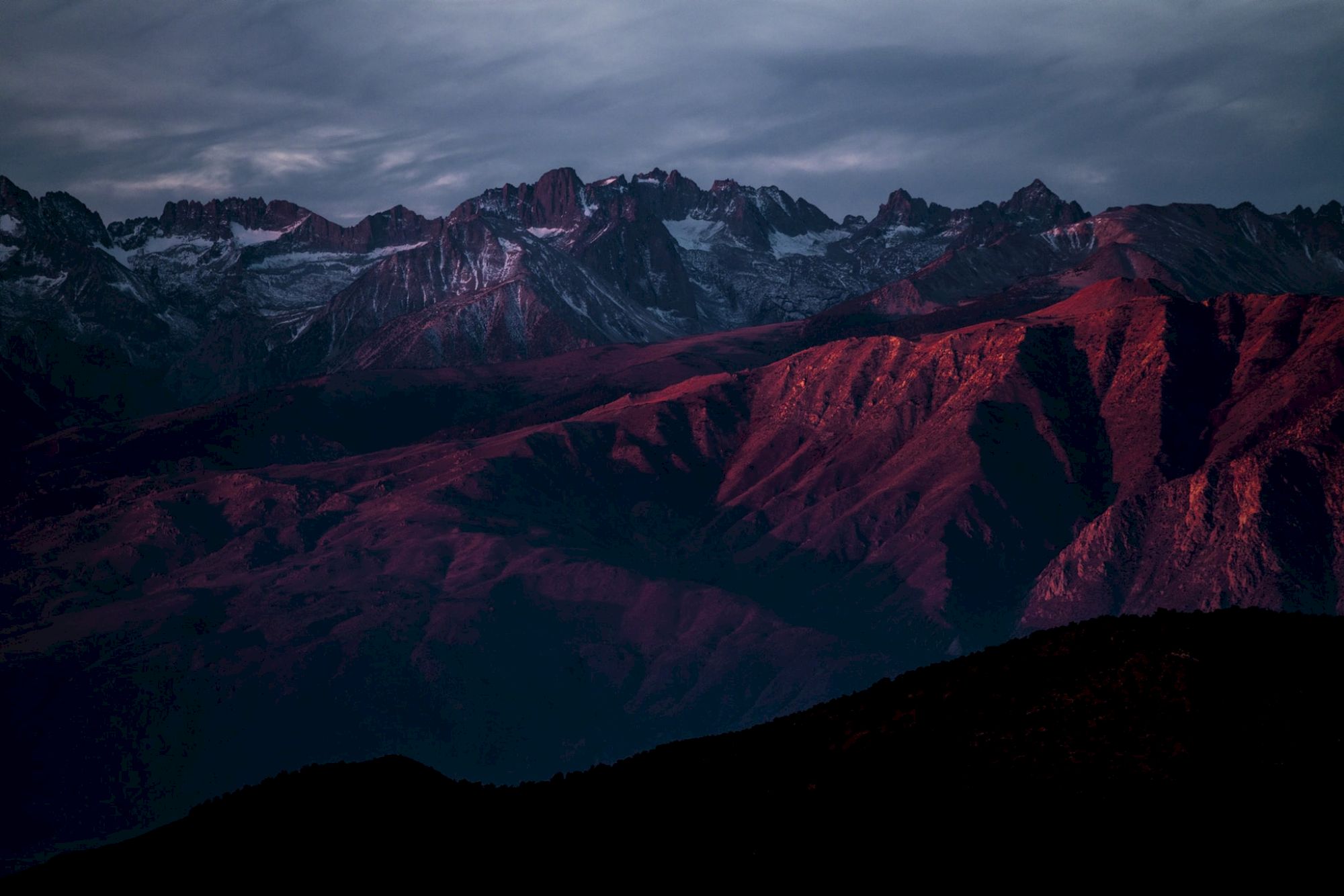 A mountain range bathed in red light under a cloudy sky during sunset or sunrise, creating a dramatic and serene scene.