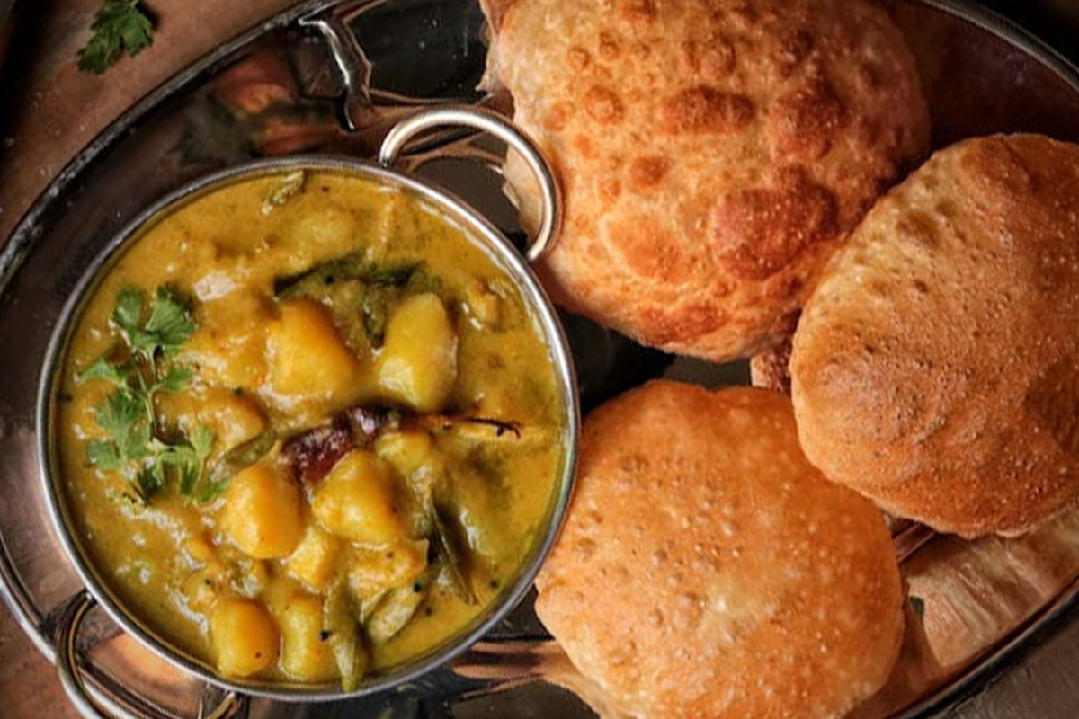 A bowl of curry with vegetables and herbs, served with three pieces of deep-fried bread on a metal tray.