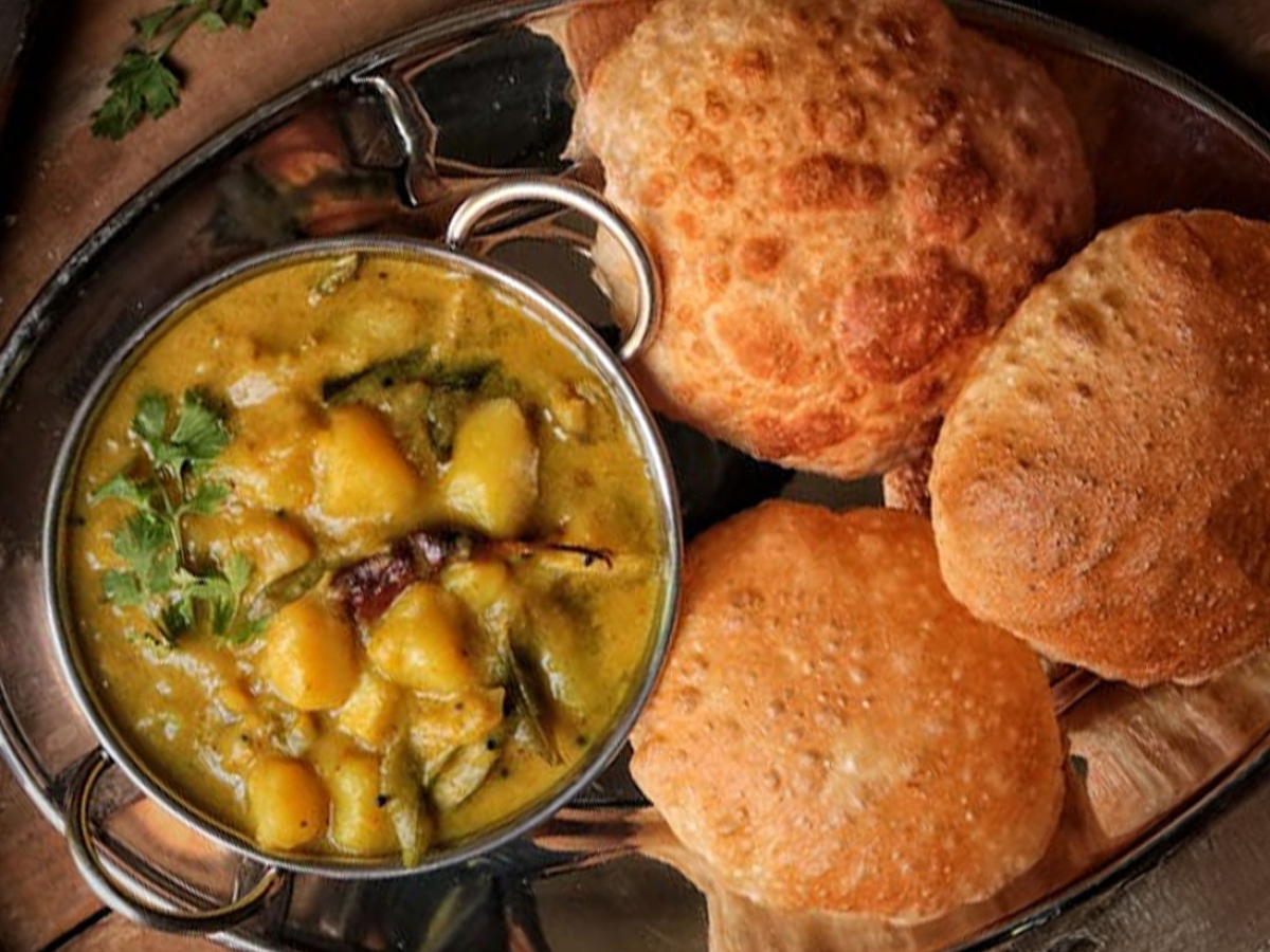 A bowl of curry with vegetables and herbs, served with three pieces of deep-fried bread on a metal tray.