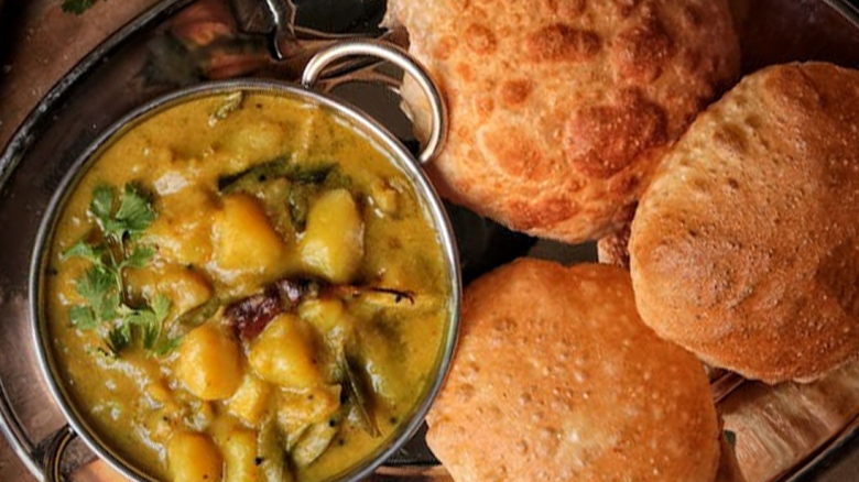A bowl of curry with vegetables and herbs, served with three pieces of deep-fried bread on a metal tray.