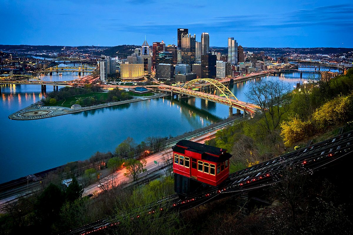 The image shows a cityscape at dusk with a yellow bridge and a red funicular, surrounded by a river and illuminated skyscrapers in the background.