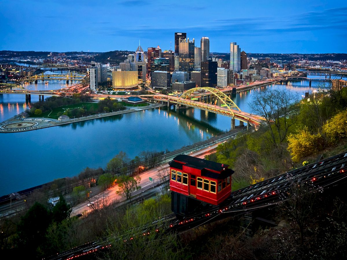 The image shows a cityscape at dusk with a yellow bridge and a red funicular, surrounded by a river and illuminated skyscrapers in the background.