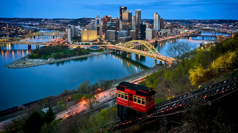 The image shows a cityscape at dusk with a yellow bridge and a red funicular, surrounded by a river and illuminated skyscrapers in the background.