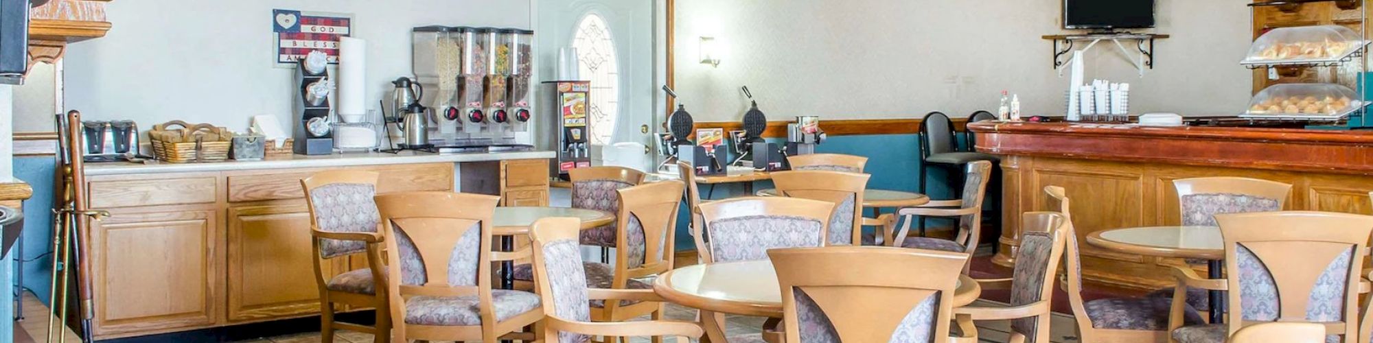 The image shows an empty cafe or dining area with wooden tables and chairs, a counter, beverage dispensers, and decorative wall plates.