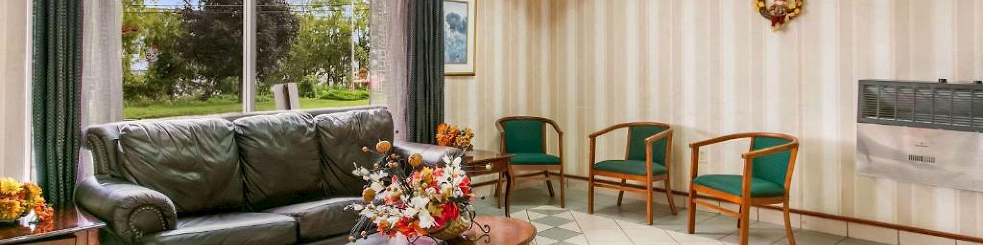 A cozy waiting room with a sofa, chairs, a clock, and floral decorations on a table, next to large windows showing trees outside.