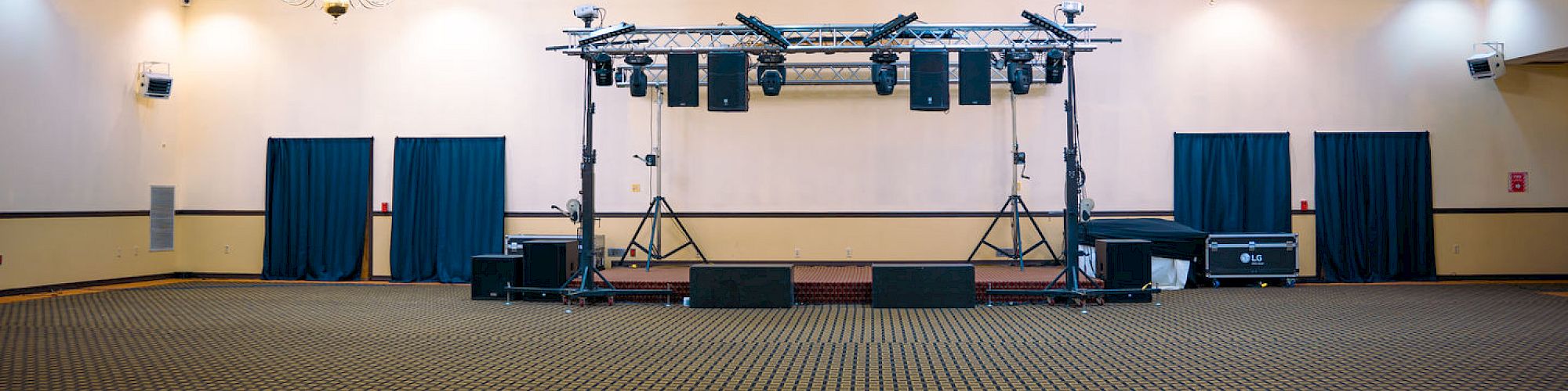 An empty banquet hall with carpeted floor, chandeliers, and a stage setup with lights and speakers in the center.