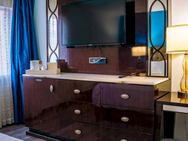 The image shows a hotel room with a TV on a wooden dresser, a lamp on a desk, and a window with curtains.