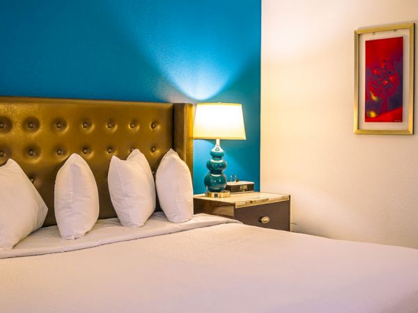 A cozy bedroom with a button-tufted headboard, pillows, a side table, lamp, and a framed artwork on the wall against a blue accent wall.