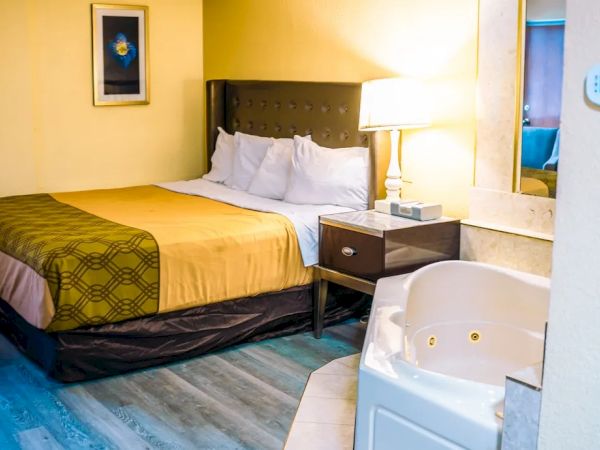 A cozy hotel room features a bed, nightstand with a lamp, wall art, and an in-room bathtub beside a wall mirror and thermostat.