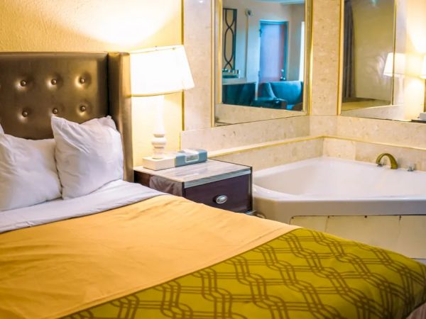 The image shows a hotel room with a bed, nightstand, lamp, and a bathtub beside it, with mirrors on the wall.