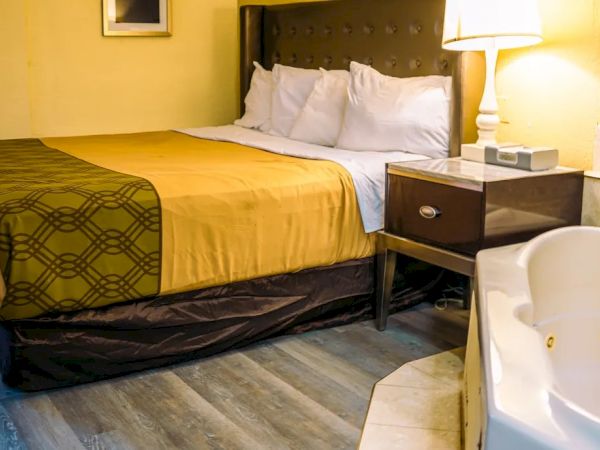 The image shows a cozy hotel room with a bed, yellow bedding, a nightstand with a lamp, and a jacuzzi tub nearby.